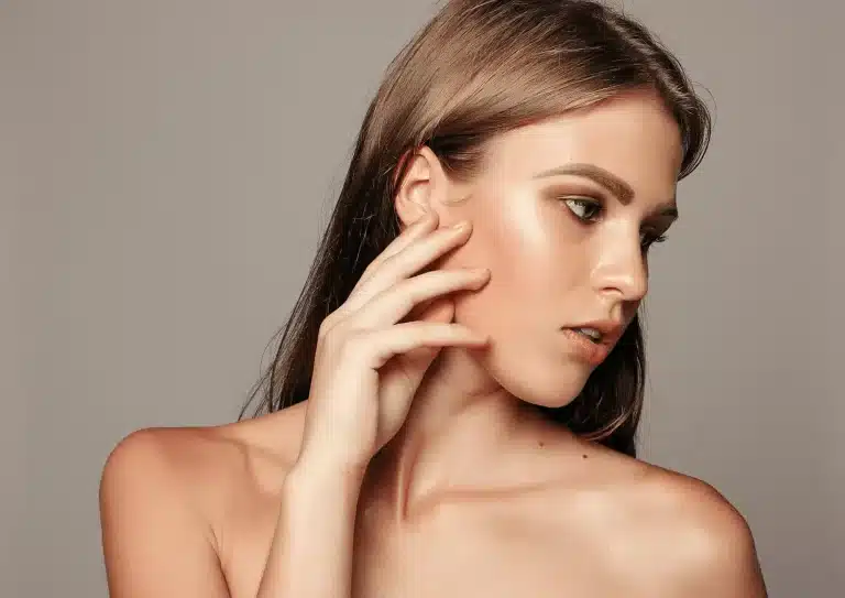 A woman with long brown hair and natural makeup poses against a neutral background. She touches her face lightly with one hand, showcasing a contemplative and serene expression with her gaze directed to the side. Her shoulders are exposed.
