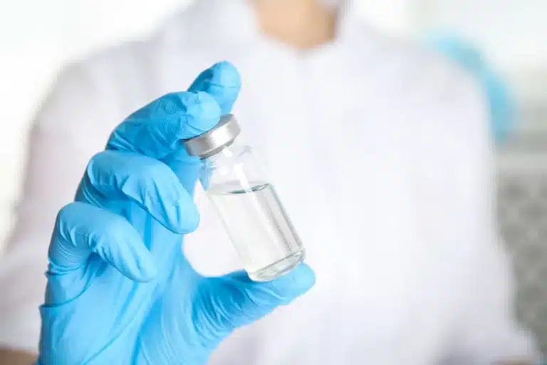 A person wearing blue gloves holds a small glass vial containing a clear liquid. The person is dressed in a white lab coat, suggesting a medical or laboratory setting.