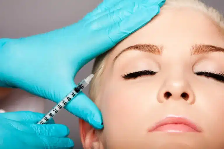 A person with closed eyes undergoes a cosmetic procedure. A medical professional wearing blue gloves holds a syringe close to their forehead, preparing for injection. The person's face is calm and relaxed, and the focus is on the injection process.