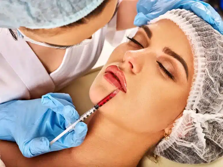 A woman, wearing a hairnet, is lying down with closed eyes. A professional, also in a hairnet and gloves, is seen administering an injectable treatment to her lips using a syringe filled with a red substance. The setting appears to be a medical or cosmetic clinic.