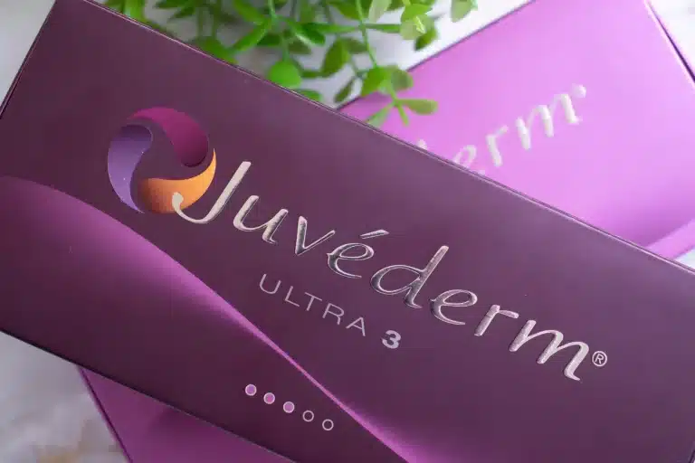 A close-up of a Juvederm Ultra 3 product box. The box is dark purple with a white and purple logo and the brand name "Juvéderm" written in a silver, cursive font. There is a green plant partially visible in the background.