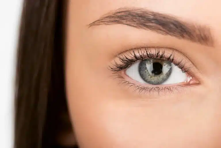 Close-up image of a human eye with greenish-blue iris and well-defined eyelashes. The eye is framed by a neatly shaped eyebrow and smooth, light skin. The image focuses solely on one eye, filling the majority of the frame.