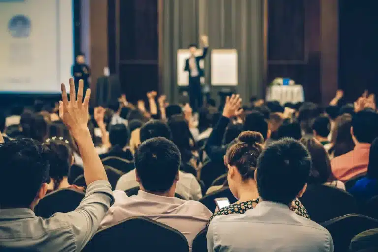 A speaker stands at the front of a conference room at AMWC 2023, presenting to an engaged audience. Many attendees are seated, with a few raising their hands, possibly to ask questions or participate. A projector screen and presentation materials are visible in the background.