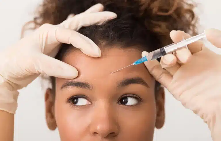 At AMWC 2023, a woman with curly hair receives an injection in her forehead. The hands of the person administering the injection, wearing white gloves, hold her skin taut as the needle approaches. The background is plain and light-colored.