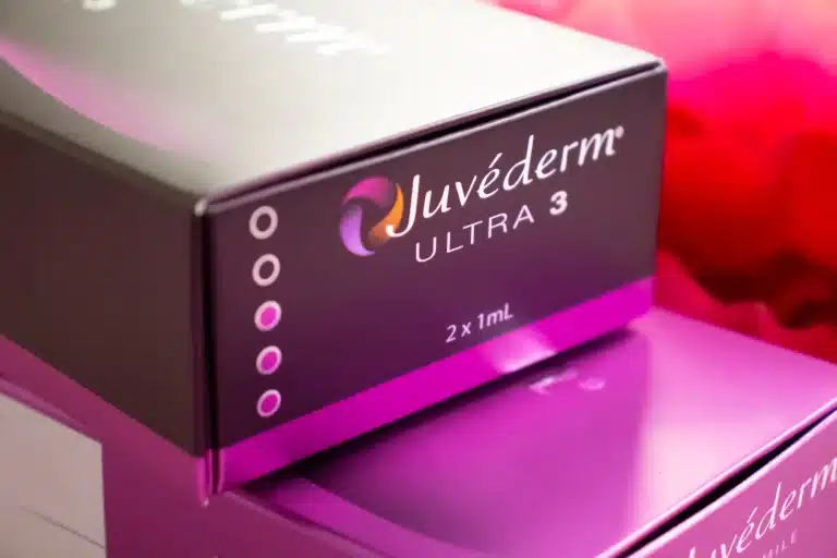 A close-up of a purple and black box labeled "Juvéderm Ultra 3" with "2 x 1mL" written underneath. The box features a sleek design with a gradient color scheme and multiple circles on the side, positioned against a blurred pink-red background reminiscent of AMWC 2023 aesthetics.