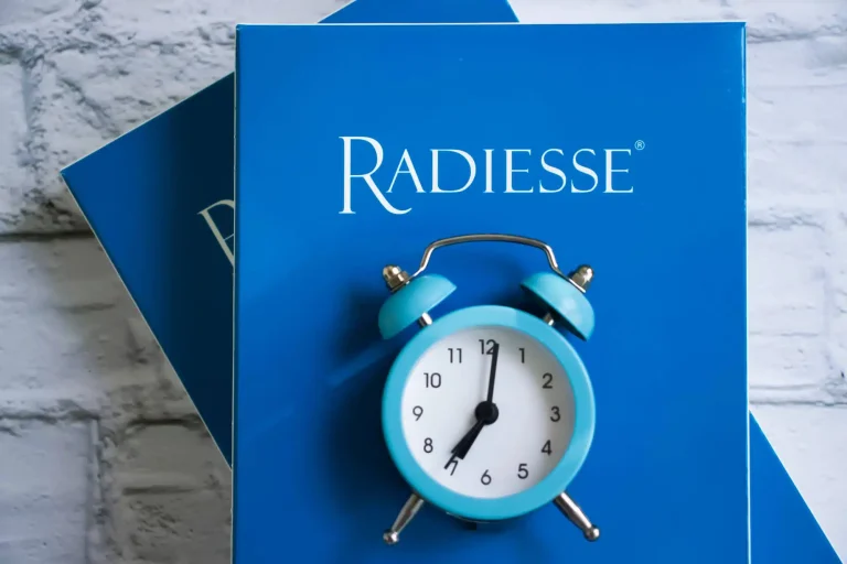 A small blue alarm clock, displaying two o'clock, is placed on top of a blue box labeled "Radiesse." The background features a textured white surface, reminiscent of the elegant settings at AMWC 2023.