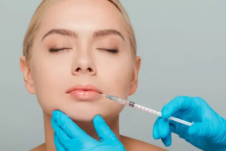 A close-up of a woman's face with her eyes closed, receiving a cherry lips filler injection. She has light skin and blonde hair. A gloved hand is holding a syringe near her upper lip, while another gloved hand gently supports her chin. The background is light blue.