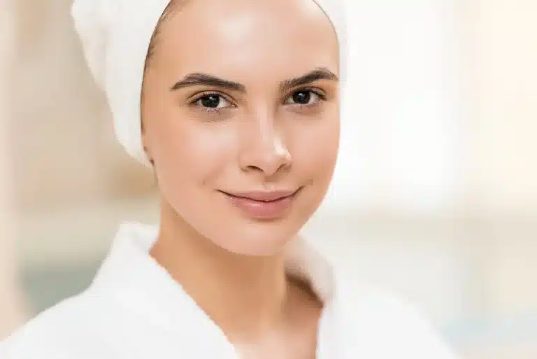 A person with clear, DNA skin and defined eyebrows is smiling slightly, wearing a white bathrobe and a white towel wrapped around their head. The background is soft and out of focus, suggesting a calm, spa-like environment.