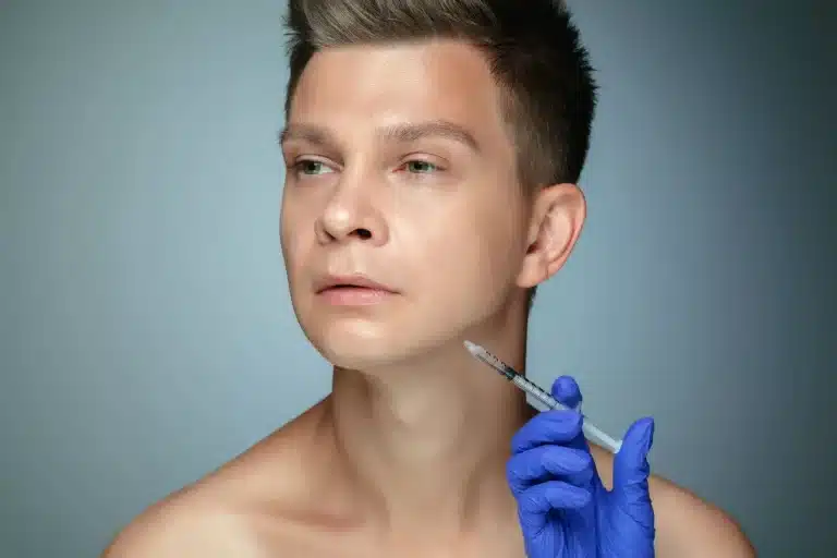 A young man with short hair is looking off to the side with a neutral expression. A gloved hand is holding a syringe near his chin, suggesting a cosmetic procedure. The background is neutral, emphasizing the focus on the man's face and the syringe, as if enhancing his DNA for flawless skin.