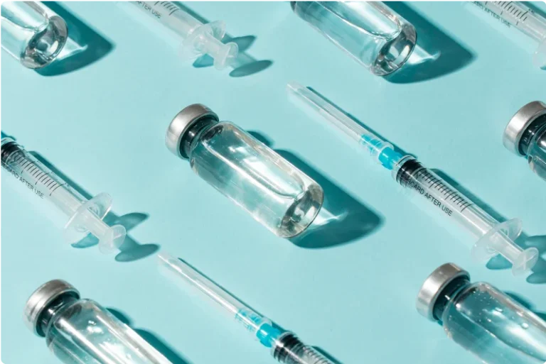 A flat lay photo of multiple clear glass vials with metal lids and several syringes on a light blue background. The vials and syringes, perhaps for Juvederm Ultra vs Ultra Plus treatments, are neatly arranged in a diagonal pattern, creating a clean and organized composition.