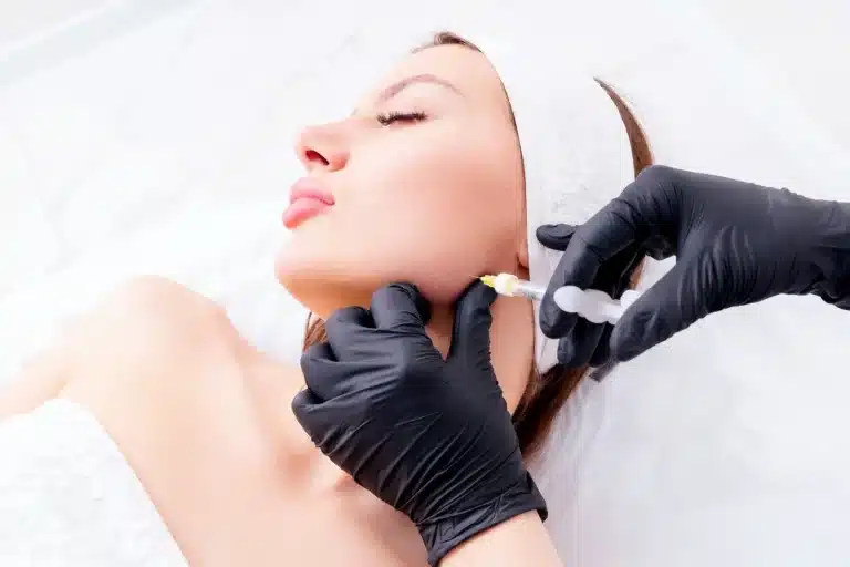 A woman lying down with her eyes closed is receiving Belkyra injections in her jaw area from a professional wearing black gloves. The woman has a white headband and towel. The background is bright and clinical.