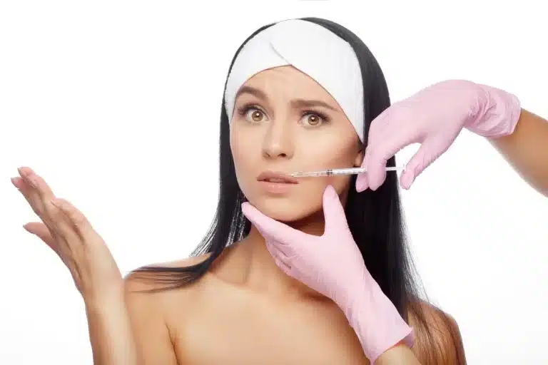 A woman with long dark hair, wearing a white headband, looks uncertain as she holds her left hand up. Another person in pink gloves is holding her face and administering Belkyra injections to her cheek with a syringe. The background is white.