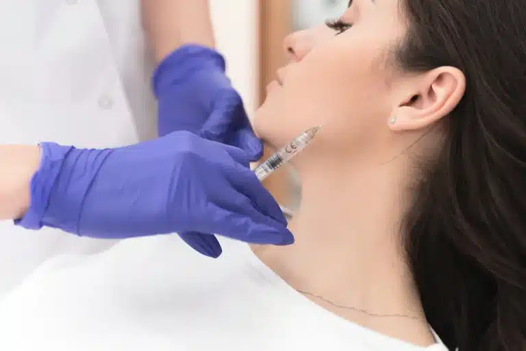 A person with dark hair is leaning back while a gloved hand holds a syringe near their jawline, preparing for an injection. The person appears calm, and the environment is clinical, suggesting a medical or cosmetic procedure.