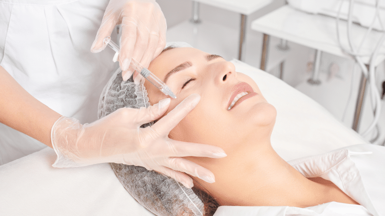 A person lies on a treatment bed with eyes closed while a clinician administers a Belotero filler injection under the eyes. The clinician wears gloves and protective clothing, and the patient dons a hair cover. The setting appears to be a medical or cosmetic clinic.