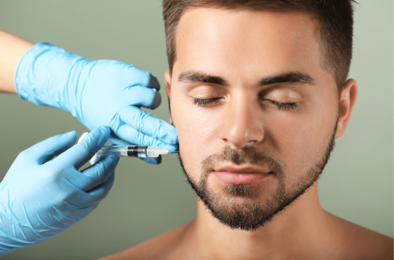 A person with short hair and a beard is receiving a Revanesse injection on the side of their face. The individual administering the treatment is wearing blue gloves. The background is a plain, muted green color, setting the stage for an intriguing before and after transformation.