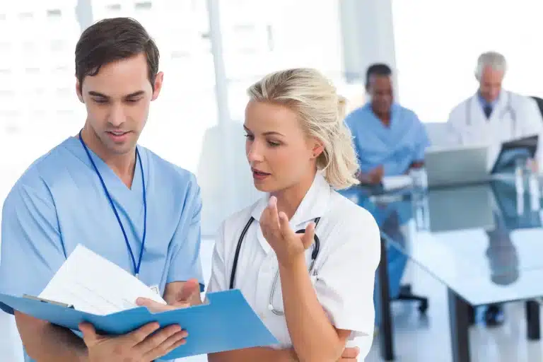 Two medical professionals, one man in light blue scrubs and one woman in a white lab coat, discuss a patient's file while three other healthcare workers are in the background, focused on their tasks. The setting appears to be a modern medical office or hospital.