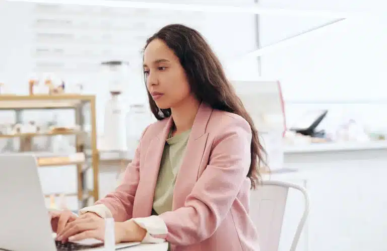 A person with long dark hair is sitting at a table in a bright, modern cafe, typing on a laptop. They are wearing a pink blazer over a light green top and appear focused on their work. The background includes shelves with various items and a blurred coffee machine.