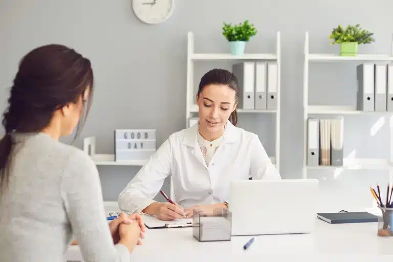 A doctor in a white coat sits at a desk with a laptop, writing notes while talking to a seated female patient. The office is modern with organized shelves, a clock on the wall, potted plants, and a sign that reads "GOOD DAY".