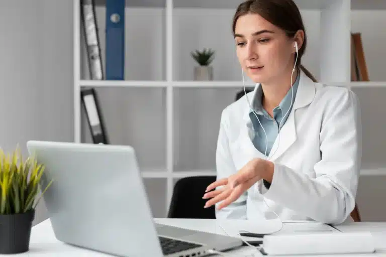 A woman in a white coat, likely a doctor, is sitting at a desk with a laptop, wearing earphones, and engaged in a video call. Behind her, there are shelves with binders and a plant. The setting appears to be a professional office.