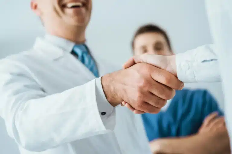 A doctor in a white coat and a striped tie shakes hands with another person while smiling. A second individual in scrubs stands in the background, slightly out of focus. The scene suggests a positive interaction in a medical setting.