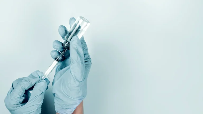 Hands in blue gloves holding a syringe and vial against a light blue background.