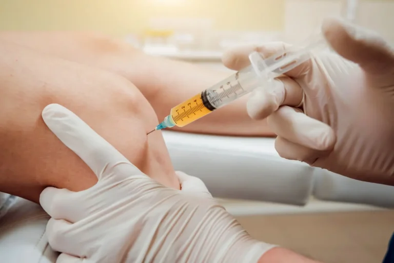 A person receiving an injection in the knee by a healthcare professional wearing gloves. The syringe contains a yellow liquid. The patient is seated on a white medical bed, and the clinical setting is neutral and well-lit.