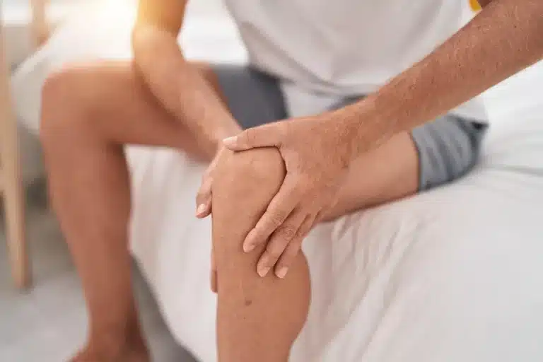 A person sitting on a bed, holding their knee with both hands, possibly indicating knee pain or discomfort. The person is wearing a light-colored shirt and shorts, and the background is softly lit.