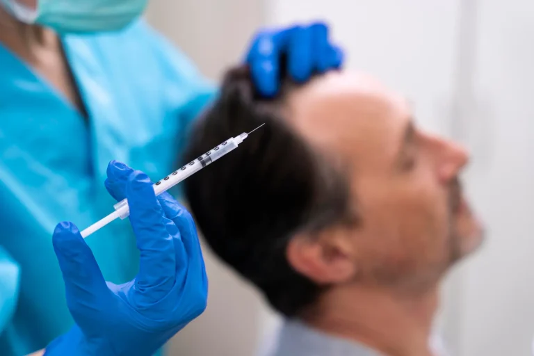 A healthcare professional in blue scrubs and gloves holds a syringe near a man's head. The man is seated, eyes closed, with a hand on his hair, suggesting a medical or cosmetic procedure.