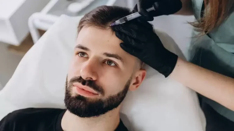 A person with a beard lies on a medical chair, receiving a scalp injection from a professional wearing black gloves.