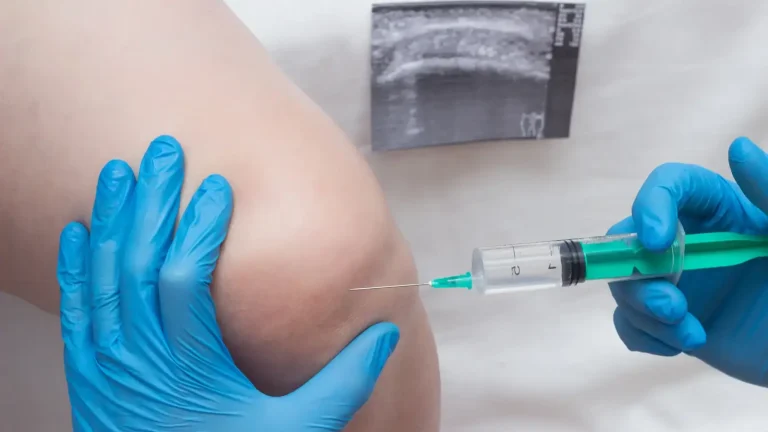 A medical professional wearing blue gloves prepares to inject a syringe into a person's knee. An ultrasound image is visible in the background, suggesting a medical procedure or treatment underway.