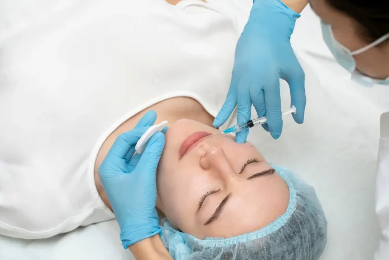 A person lying down receives a facial injection from a healthcare professional wearing blue gloves and a mask. The person has a hair cover and closed eyes, suggesting a medical or cosmetic procedure.