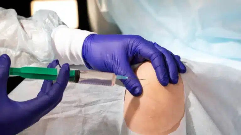 A medical professional wearing blue gloves administers an injection into a patient's knee in a clinical setting. The patient is draped with a sterile sheet to ensure a clean procedure.