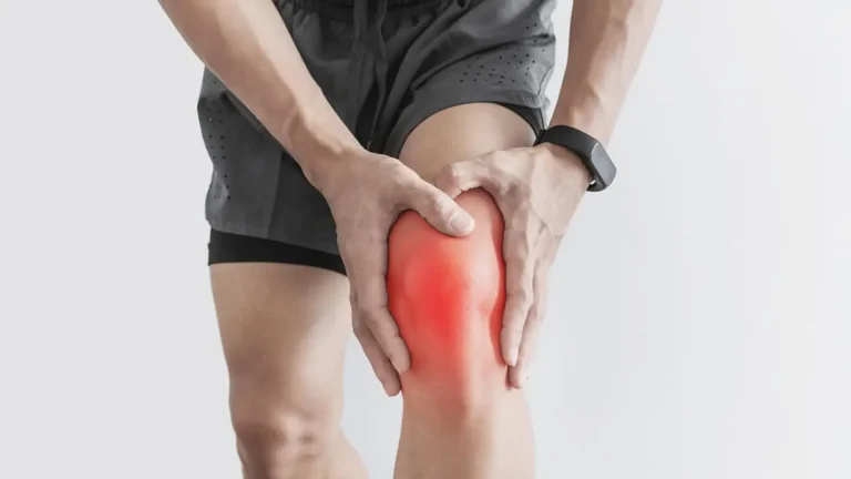 A person in sportswear holding their knee, which appears red, indicating pain or injury. They are wearing a black smartwatch on their left wrist. The background is plain white.