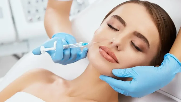 A person receives a cosmetic injection in the cheek while lying down. They have light brown hair and wear a white towel. A professional wearing blue gloves administers the procedure.