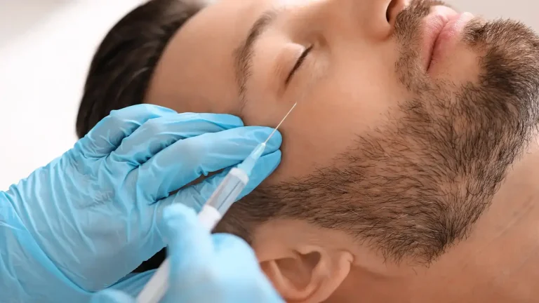 A person with a beard lies down with eyes closed while a gloved hand administers an injection to the cheek using a syringe.