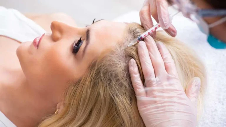 A person with blonde hair lies on a white surface while receiving an injection in the scalp. A practitioner wearing gloves administers the treatment. The scene suggests a medical or cosmetic procedure.