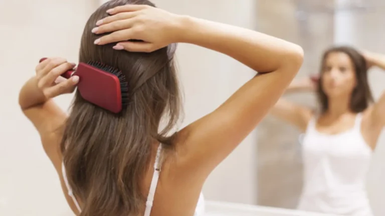 A woman is brushing her long brown hair with a red hairbrush, standing in front of a mirror. She is wearing a white tank top and is focused on grooming her hair. The background is softly blurred.