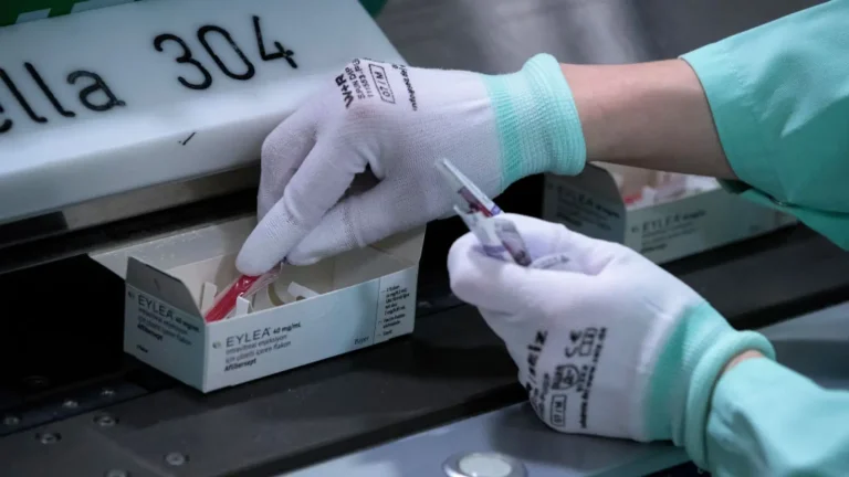 A person wearing gloves inspects boxes labeled "EYLEA" on a conveyor belt. They hold a syringe in one hand while checking the contents of an open box with the other. The background shows part of a machine with the number 304.