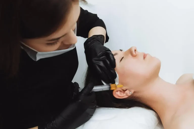 A person lying on a bed receives a facial injection from a professional wearing black gloves. The professional is focused and leans over the patient in a clinical setting with white walls.