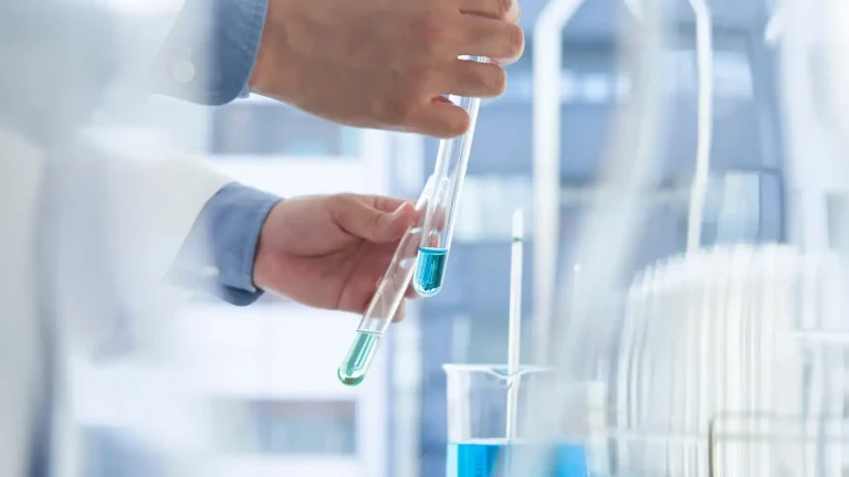 A person in a lab coat holds two test tubes with blue liquid, conducting an experiment. A beaker with blue liquid is placed on the table. The background is a laboratory setting with blurred equipment visible.