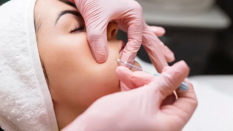 A person lying down with eyes closed, wearing a white headband. They are receiving an injection near the lips from someone wearing pink gloves, using a syringe. The background is blurred.