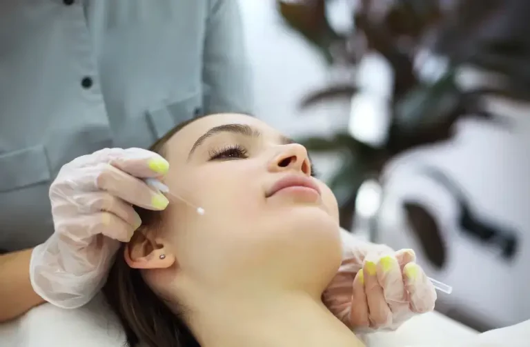 A person receives a cosmetic facial treatment from a professional wearing gloves. The professional holds a small applicator near the person's cheek in a clinic setting. The person looks relaxed with eyes closed, lying on a white surface.