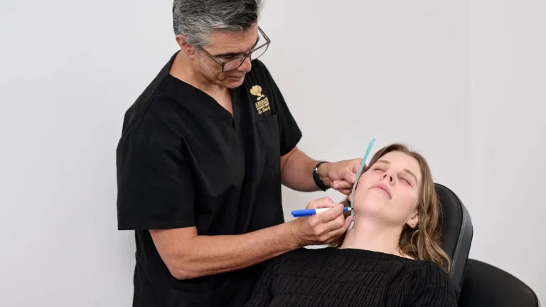 A doctor in black scrubs marks a patient's face with a blue pen. The patient, wearing a black top, is reclining in a chair with her eyes closed. They are in a clinical setting with a plain white background.