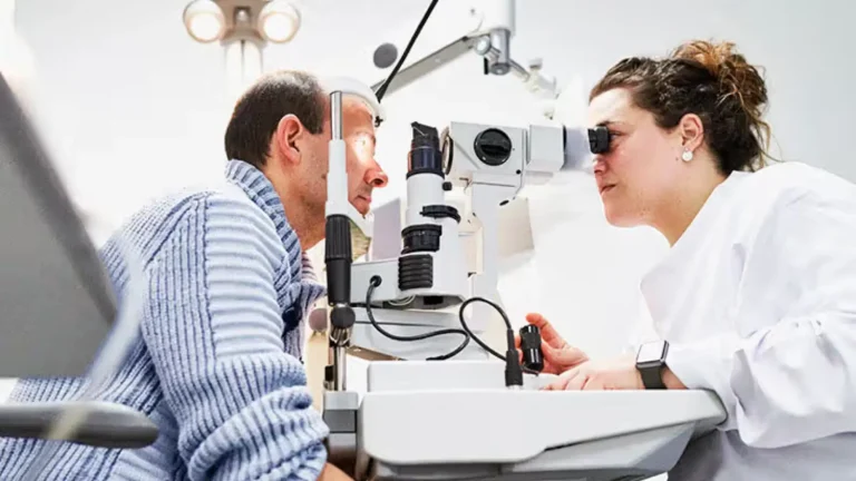 A woman in a lab coat uses a slit lamp to examine the eyes of a seated man. The equipment features multiple lenses and lights. They are in a brightly lit office, focusing on the eye examination.