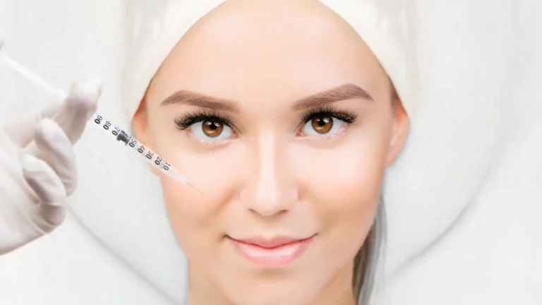 A person with smooth skin and symmetrical eyebrows is lying down. A gloved hand holds a syringe near their cheek, suggesting a cosmetic procedure. The person has a neutral expression and is wearing a hair cover.