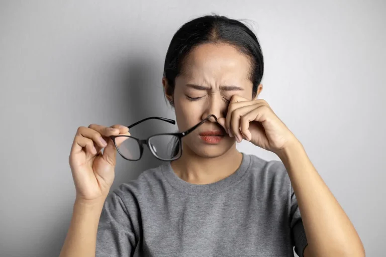 A person holding glasses in their right hand and rubbing their nose bridge with their left hand. They have a concerned expression and are wearing a grey shirt, standing against a plain grey background.