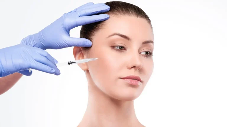 A person with tied-back hair receives a facial injection by a gloved hand holding a syringe. The background is plain white, highlighting the procedure taking place.