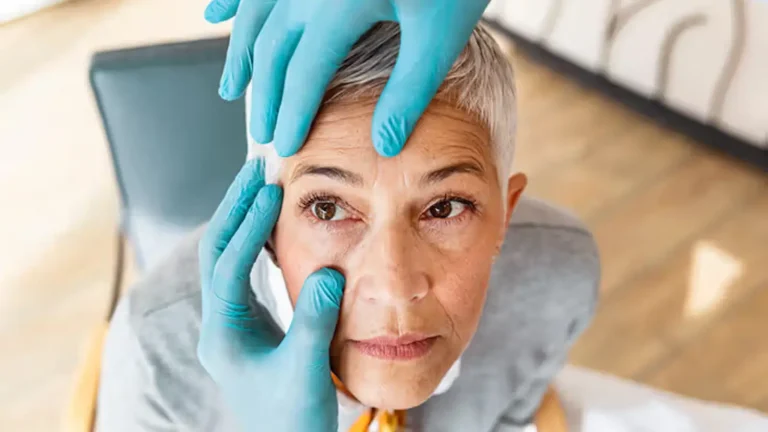 A person with short gray hair is having their eyes examined by someone wearing blue medical gloves. The person is seated and looking upwards, while the examiner gently holds their eyelids. The background is softly blurred.