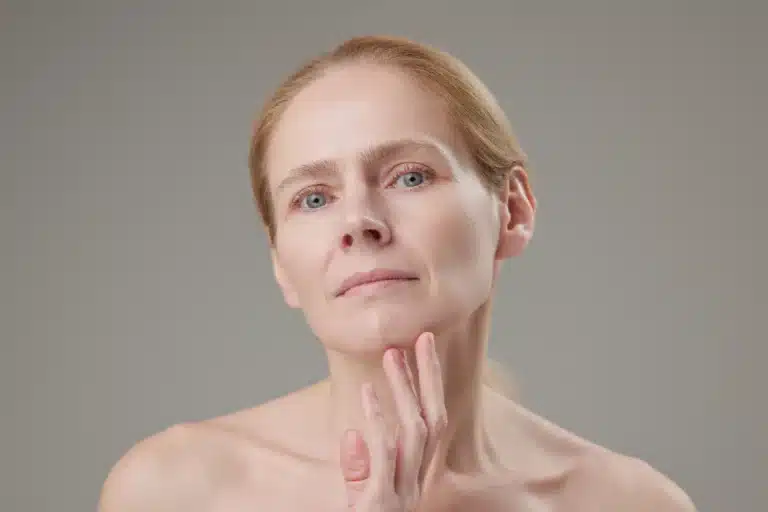 A woman with fair skin and blond hair, possibly enhanced by peptide treatments, gazes serenely into the camera. Her right hand gently touches her jawline against a neutral gray background.