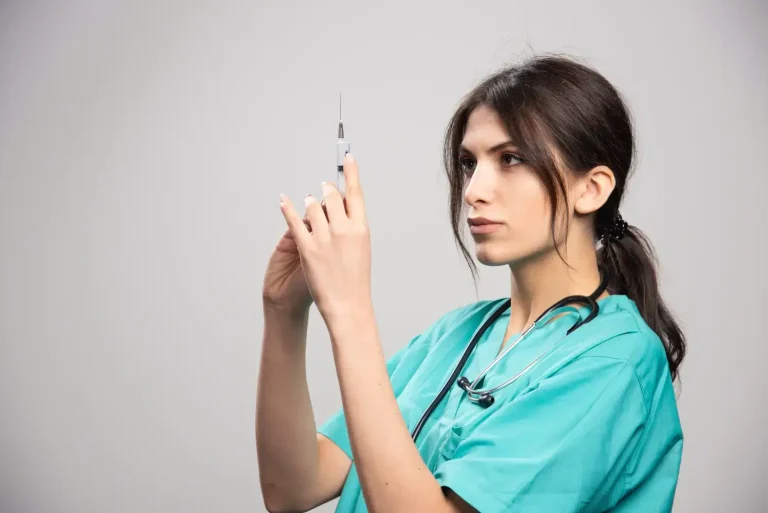 A healthcare professional wearing teal scrubs and a stethoscope prepares a syringe. They have long dark hair tied back and are focused on the syringe against a plain background.